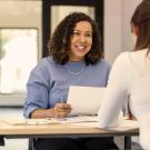 two business women sit across from each other in an office and one is holding papers